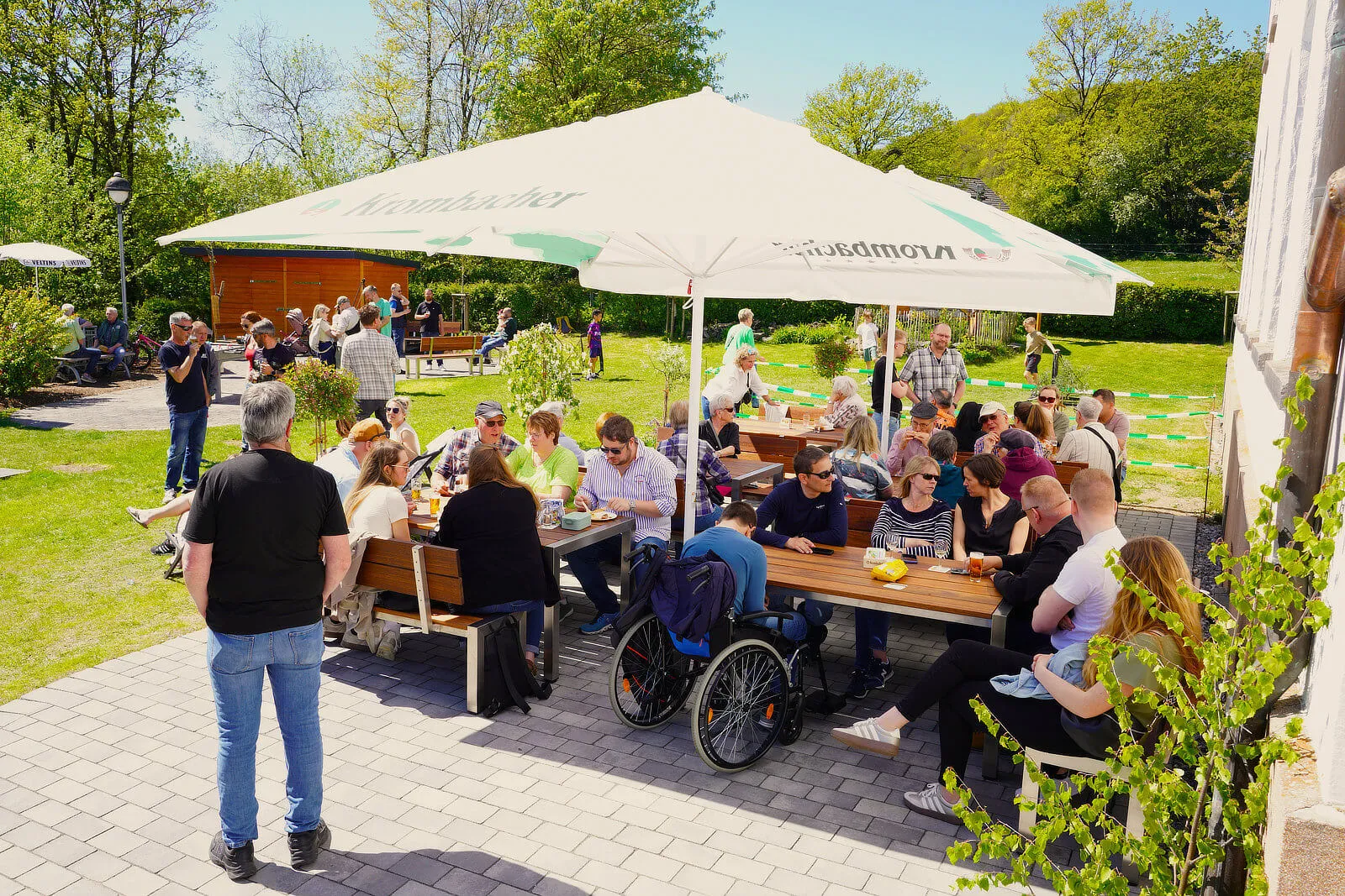 Menschen sitzen und stehen an Biertischen vor dem Burgerhaus Bremcke, einige genießen Speisen und Getränke. Im Hintergrund sind weitere Besucher und Bäume zu sehen.