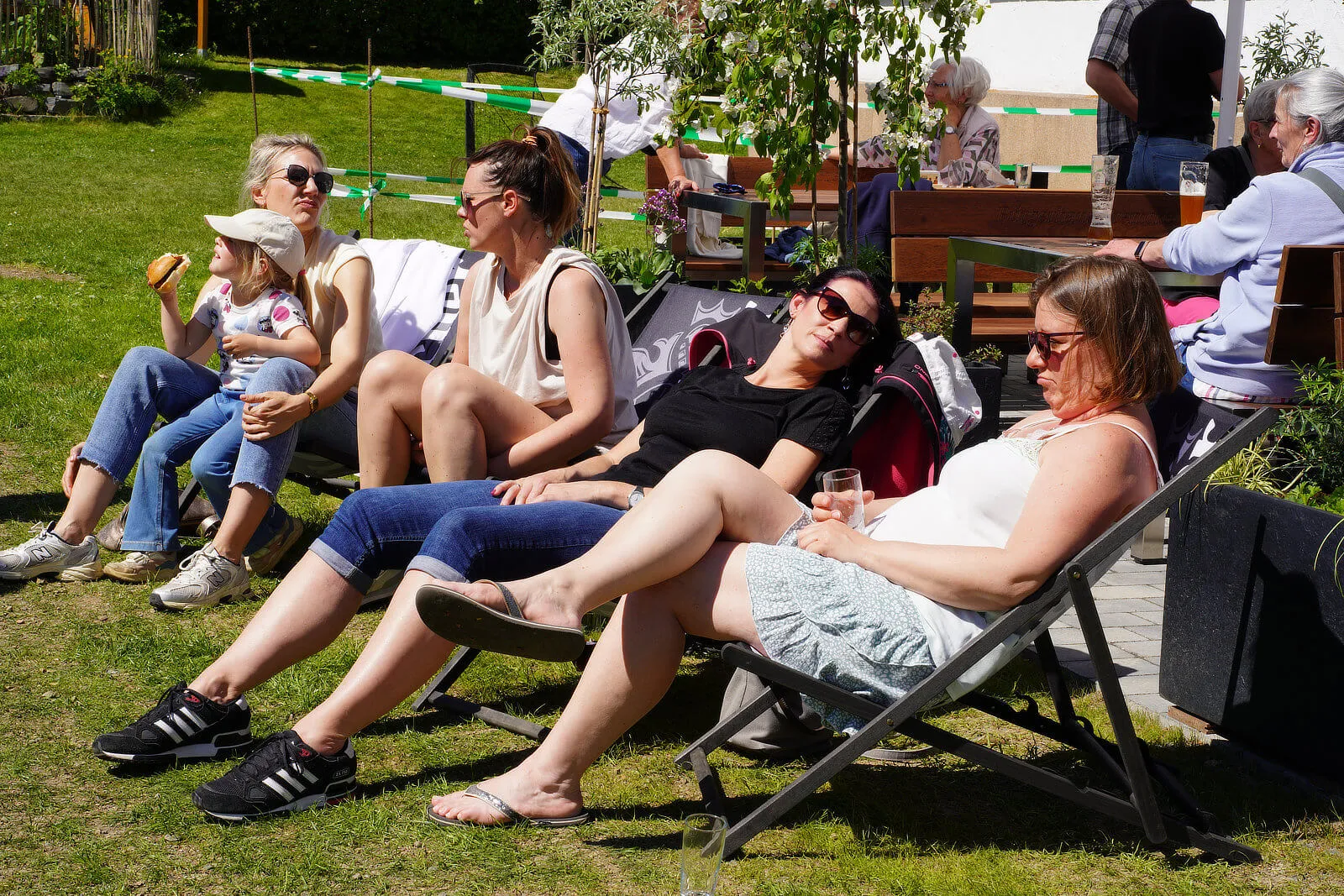 Mehrere Menschen sitzen und liegen auf einer grünen Wiese vor dem Burgerhaus Bremcke und genießen die Sonne. Einige halten Getränke oder essen.