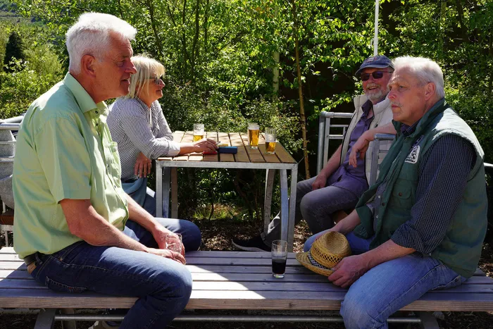 Vier ältere Menschen sitzen an einem Metalltisch im Freien, umgeben von Grün. Sie unterhalten sich und trinken Bier aus Gläsern.