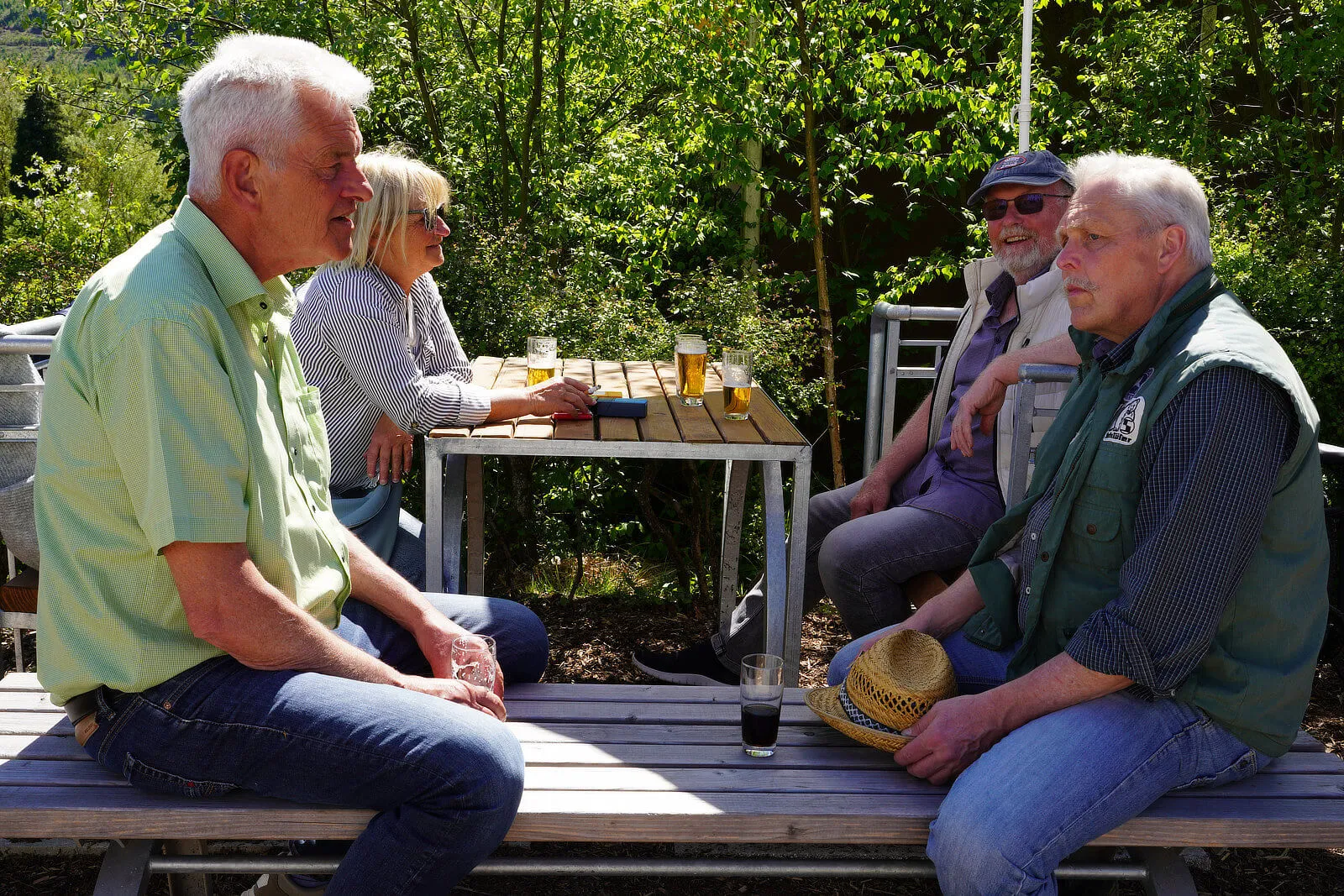 Vier ältere Menschen sitzen an einem Metalltisch im Freien, umgeben von Grün. Sie unterhalten sich und trinken Bier aus Gläsern.
