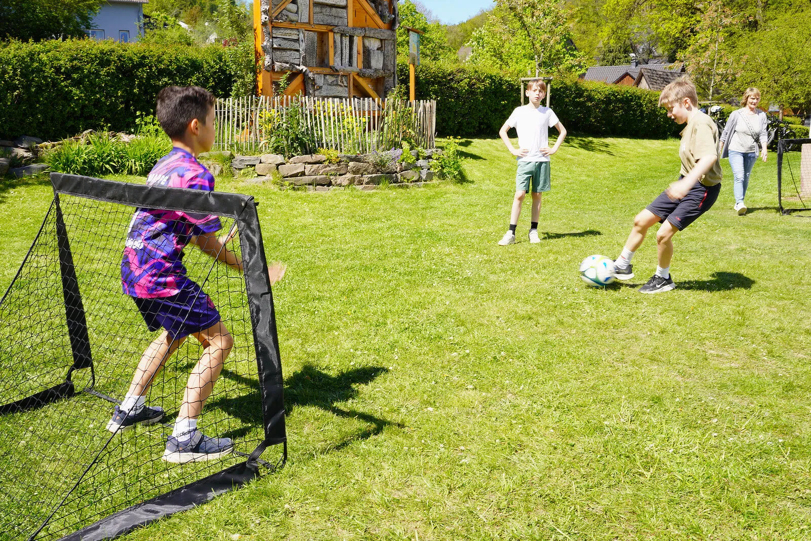 Drei Kinder spielen Fußball auf einer grünen Wiese vor einem Holzhaus. Ein Junge schießt auf das Tor, während die anderen zusehen.