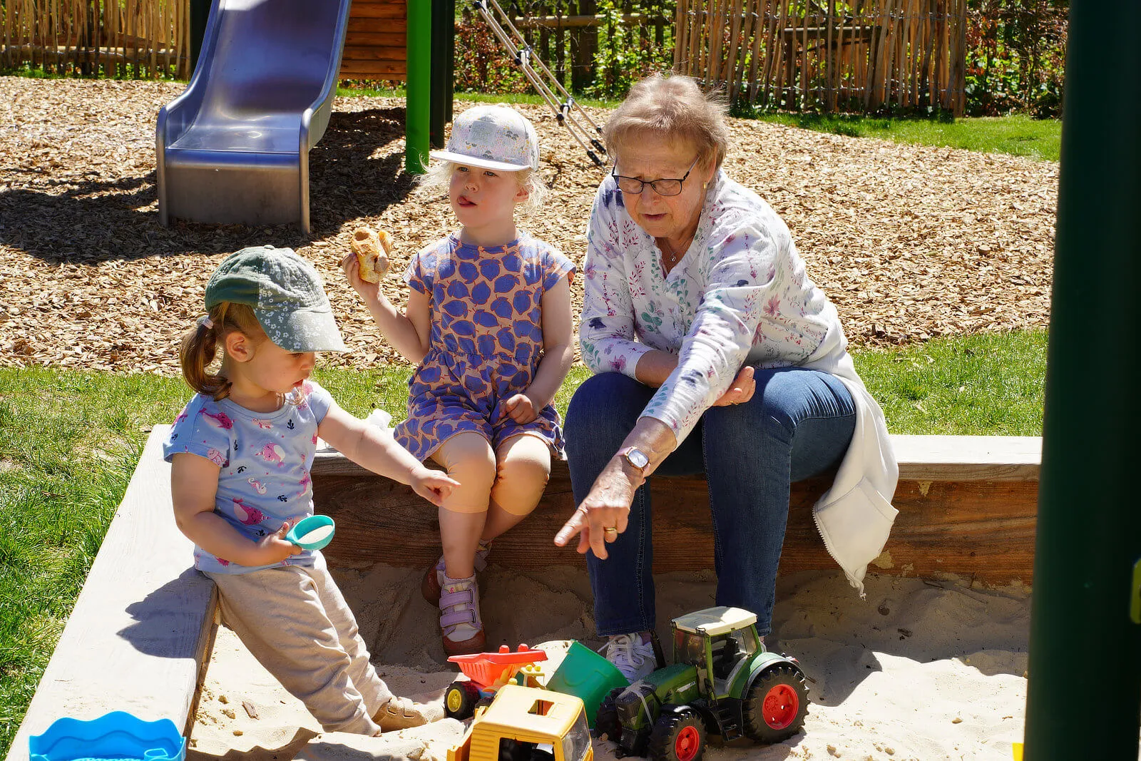 Zwei kleine Mädchen und eine ältere Frau sitzen an einem Sandkasten. Die Kinder spielen mit Sandspielzeug, während die Frau ihnen zusieht.