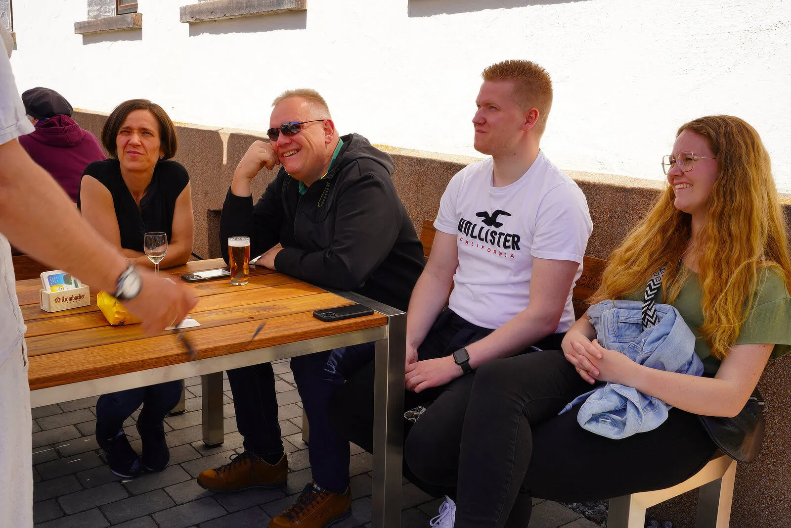 Eine Gruppe von Menschen sitzt an einem Holztisch vor einem hellen Gebäude und genießt Getränke und Gespräche im Freien.