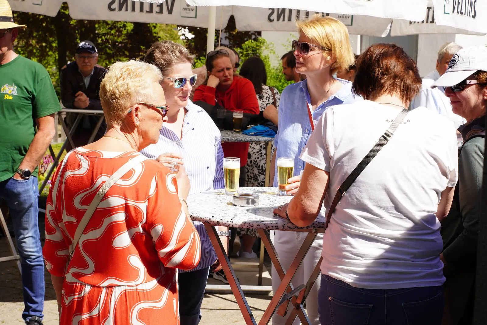 Menschen stehen und unterhalten sich an einem Stehtisch im Freien, einige halten Biergläser in der Hand. Im Hintergrund sind weitere Personen und Zelte zu sehen.