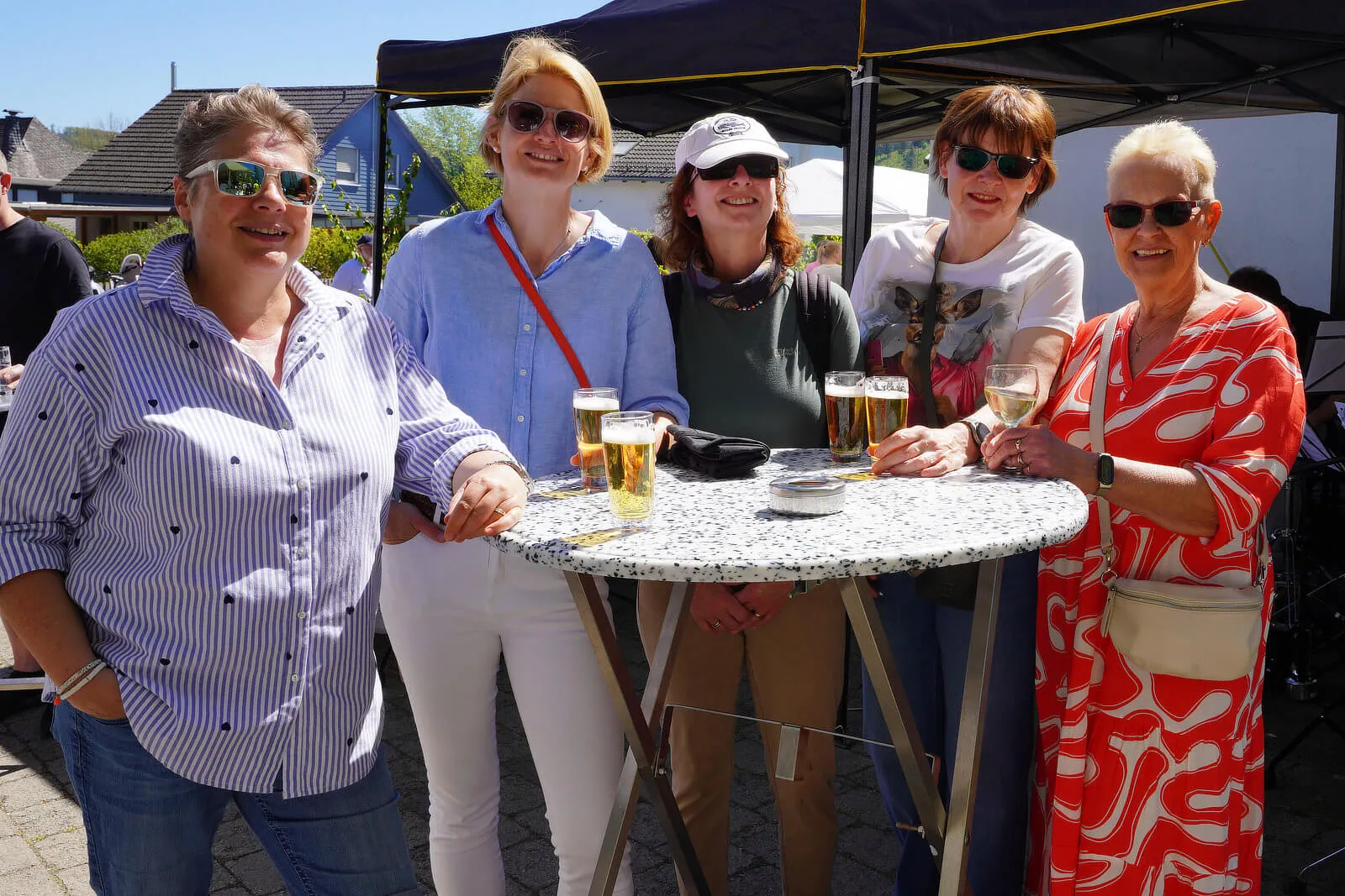 Fünf lächelnde Frauen stehen um einen runden Bistrotisch mit Biergläsern im Freien. Im Hintergrund ein Pavillon und weitere Menschen.