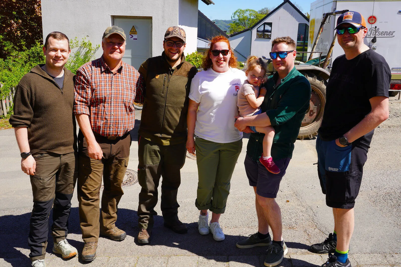 Eine Gruppe von sieben Personen steht lächelnd vor einem Gebäude mit einem Hof. Im Hintergrund sind Bäume und landwirtschaftliche Geräte zu sehen.