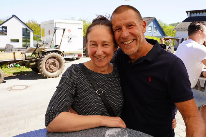 Lachendes Paar umarmt sich vor einem ländlichen Hintergrund mit Gebäuden und einem Traktor. Im Hintergrund sind weitere Menschen zu sehen.