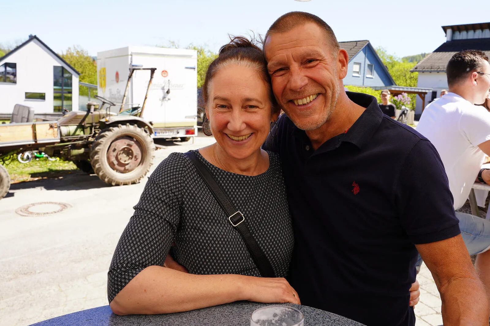 Lachendes Paar umarmt sich vor einem ländlichen Hintergrund mit Gebäuden und einem Traktor. Im Hintergrund sind weitere Menschen zu sehen.