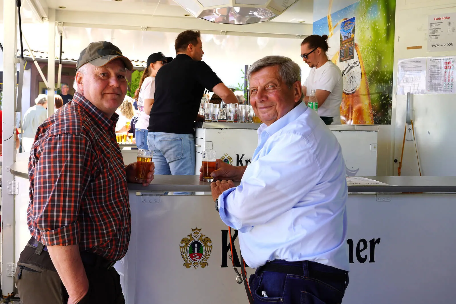 Zwei lächelnde Männer stehen an einer Bar im Freien und halten jeweils ein Glas Bier. Im Hintergrund sind weitere Menschen und ein Getränkestand zu sehen.