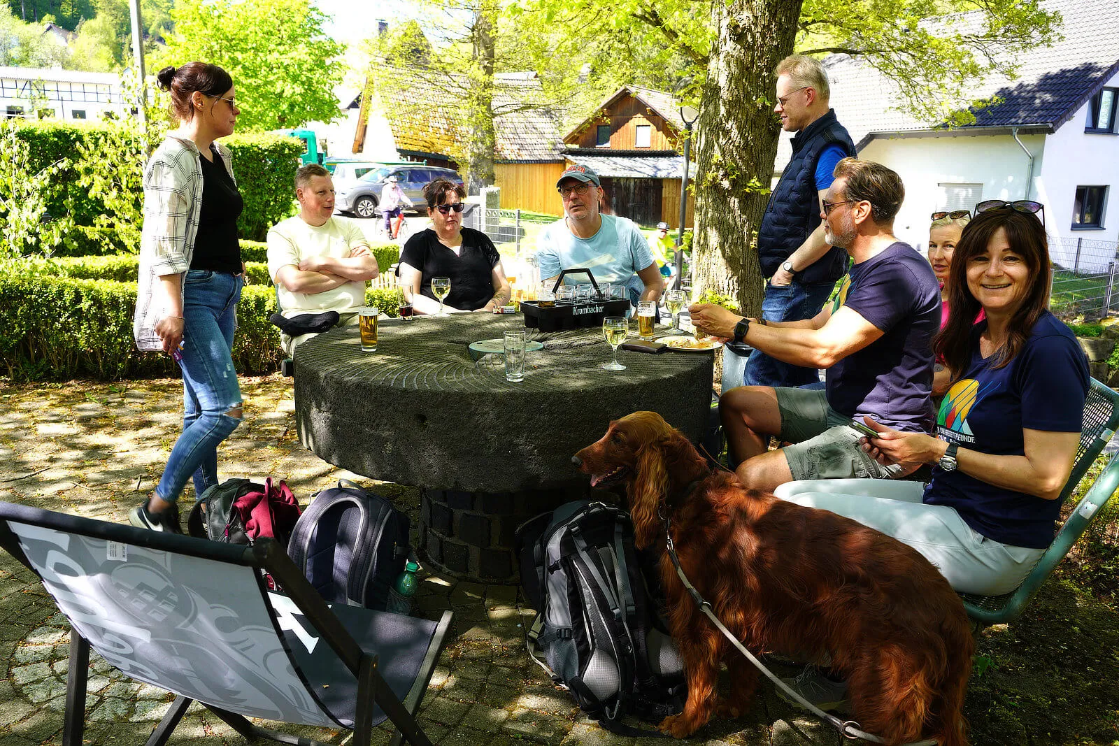 Eine Gruppe von Menschen sitzt an einem großen Steintisch vor einem traditionellen Fachwerkhaus und genießt Getränke und Gespräche. Ein brauner Hund liegt entspannt im Vordergrund, während Rucksäcke und Campingstühle daneben stehen.