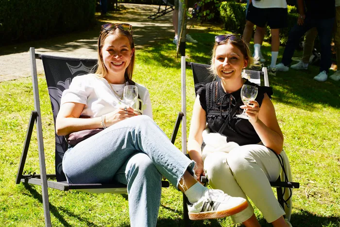 Zwei junge Frauen sitzen lächelnd auf Campingstühlen im Grünen und halten Weingläser in der Hand. Im Hintergrund sind weitere Menschen zu sehen.