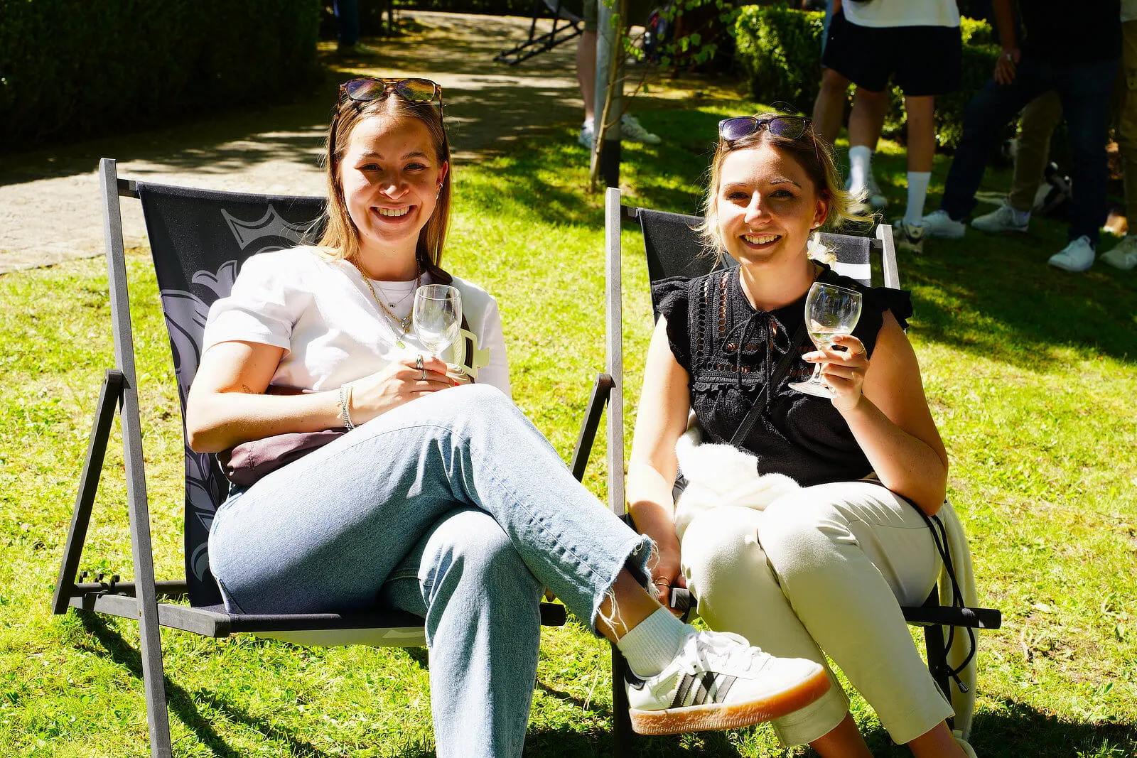 Zwei junge Frauen sitzen lächelnd auf Campingstühlen im Grünen und halten Weingläser in der Hand. Im Hintergrund sind weitere Menschen zu sehen.