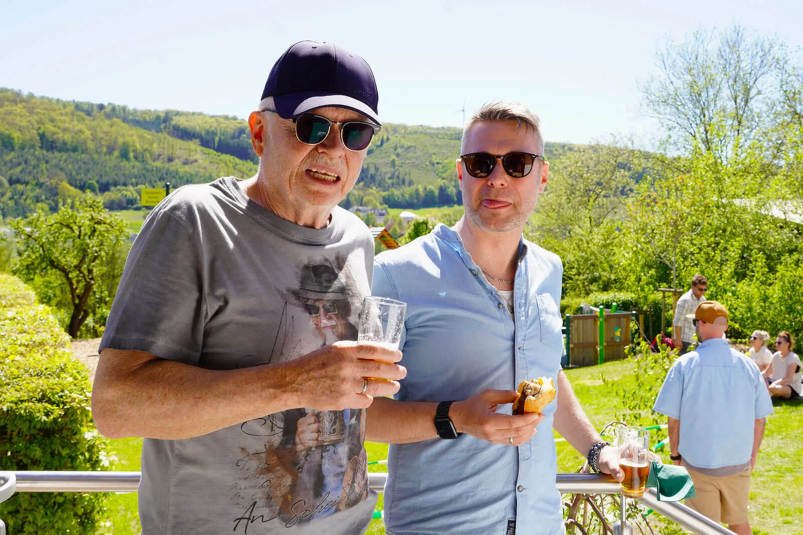 Zwei Männer, ein älterer und ein jüngerer, stehen lächelnd vor einer Geländer und halten Getränke und einen Snack in der Hand. Im Hintergrund eine grüne Hügellandschaft und weitere Menschen im Freien.