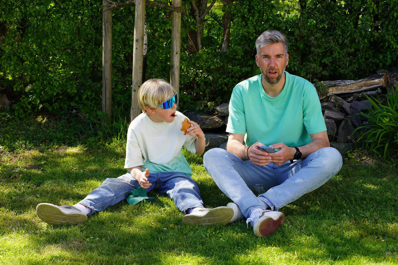 Ein Mann und ein kleiner Junge sitzen im Gras und essen. Der Junge trägt eine Sonnenbrille und ein T-Shirt, der Mann hält ein Smartphone.