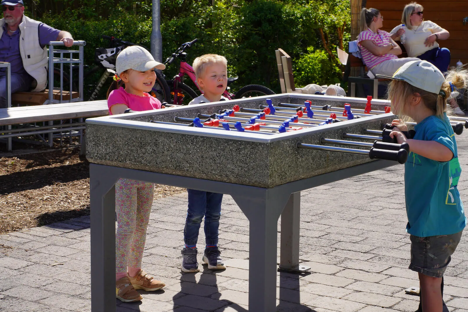 Drei Kinder spielen auf einem Kiesplatz Fußball auf einem Tischkickerspiel, während im Hintergrund Erwachsene sitzen und zusehen.