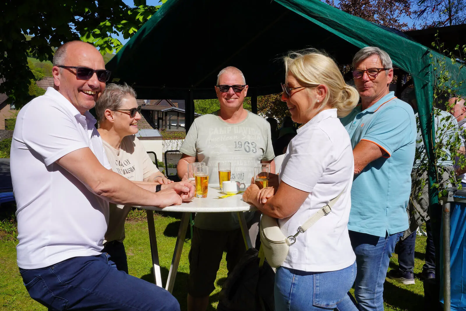 Mehrere Menschen stehen und unterhalten sich an einem Tisch mit Getränken unter einem Pavillon während eines Frühlingsfestes.