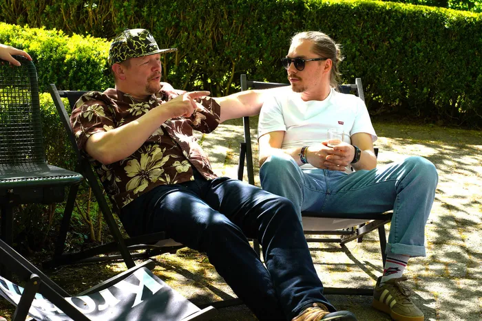 Zwei Männer sitzen im Freien auf Campingstühlen und unterhalten sich. Einer trägt eine bunte Hawaii-Hemd und eine Kappe, der andere ein weißes T-Shirt und eine Sonnenbrille.