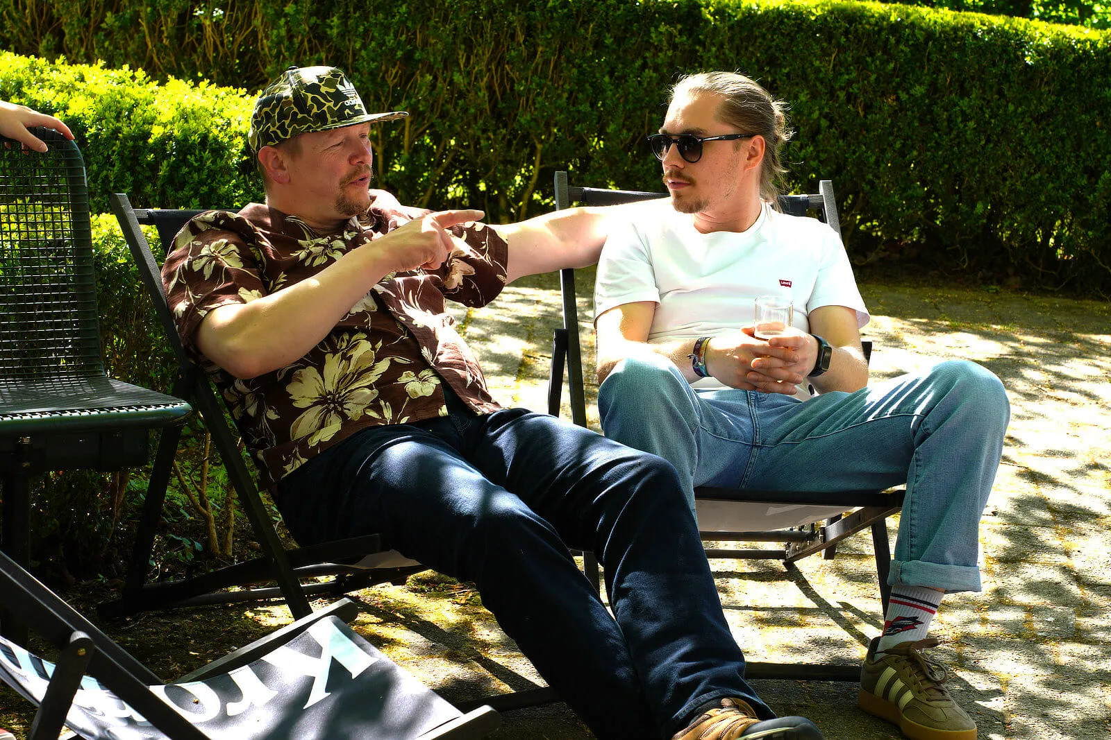 Zwei Männer sitzen im Freien auf Campingstühlen und unterhalten sich. Einer trägt eine bunte Hawaii-Hemd und eine Kappe, der andere ein weißes T-Shirt und eine Sonnenbrille.