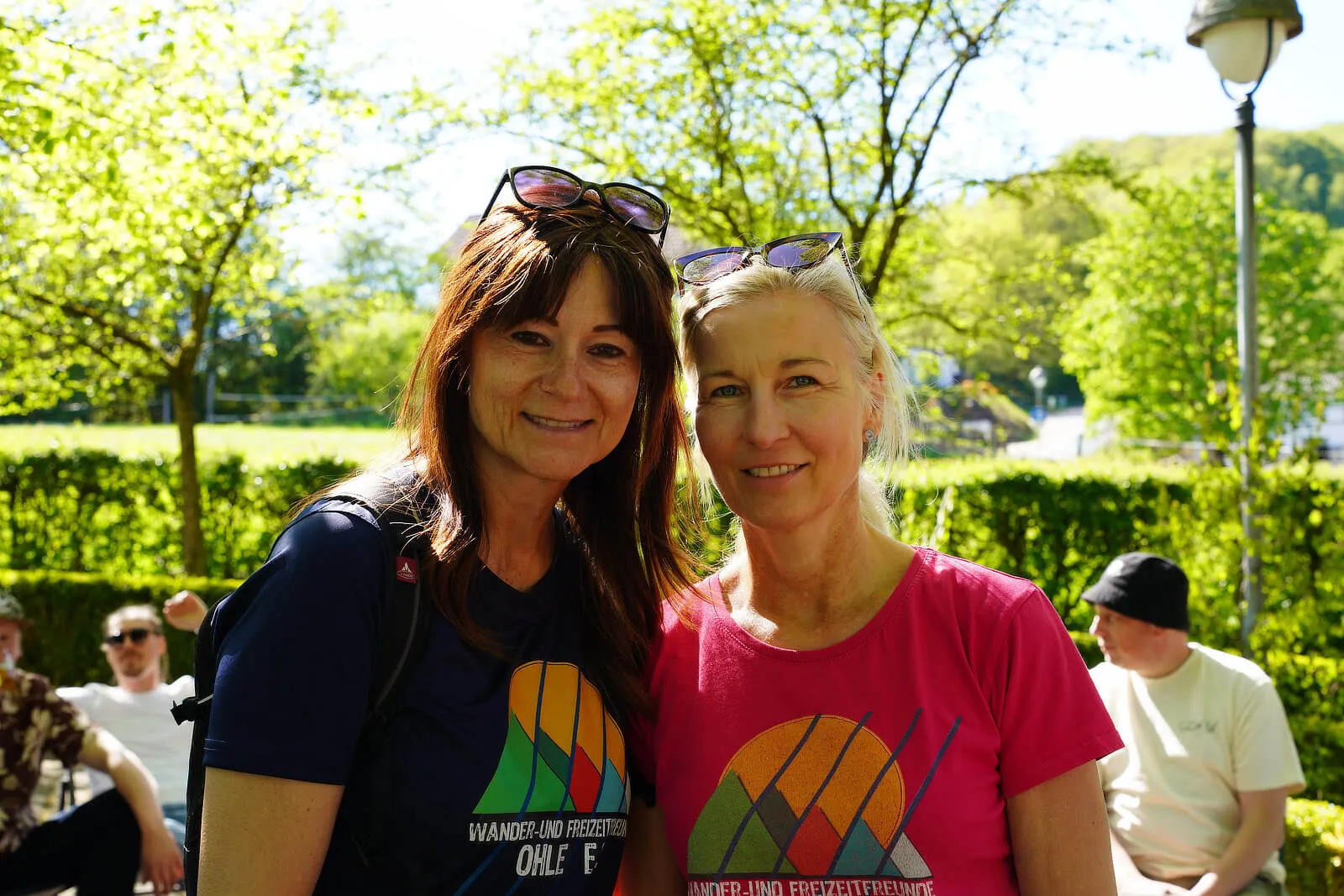 Zwei lächelnde Frauen posieren im Freien, beide tragen T-Shirts mit farbigen Streifenmotiven und die Aufschrift „Wander- und Freizeitverein Ohle“.