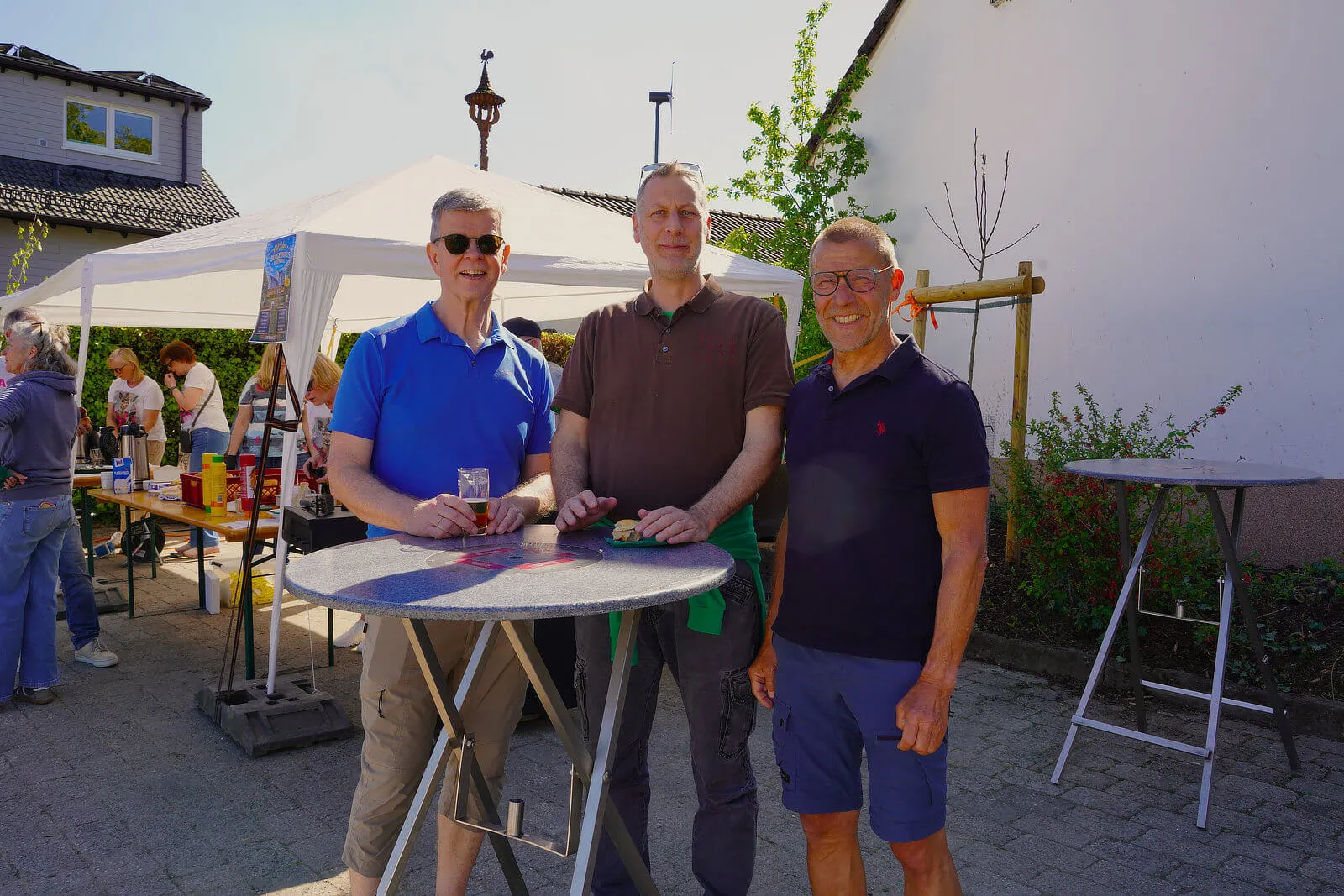Drei Männer stehen lächelnd vor einem Stehtisch im Freien, im Hintergrund ein weißes Zelt und weitere Menschen bei einem Frühlingsfest.