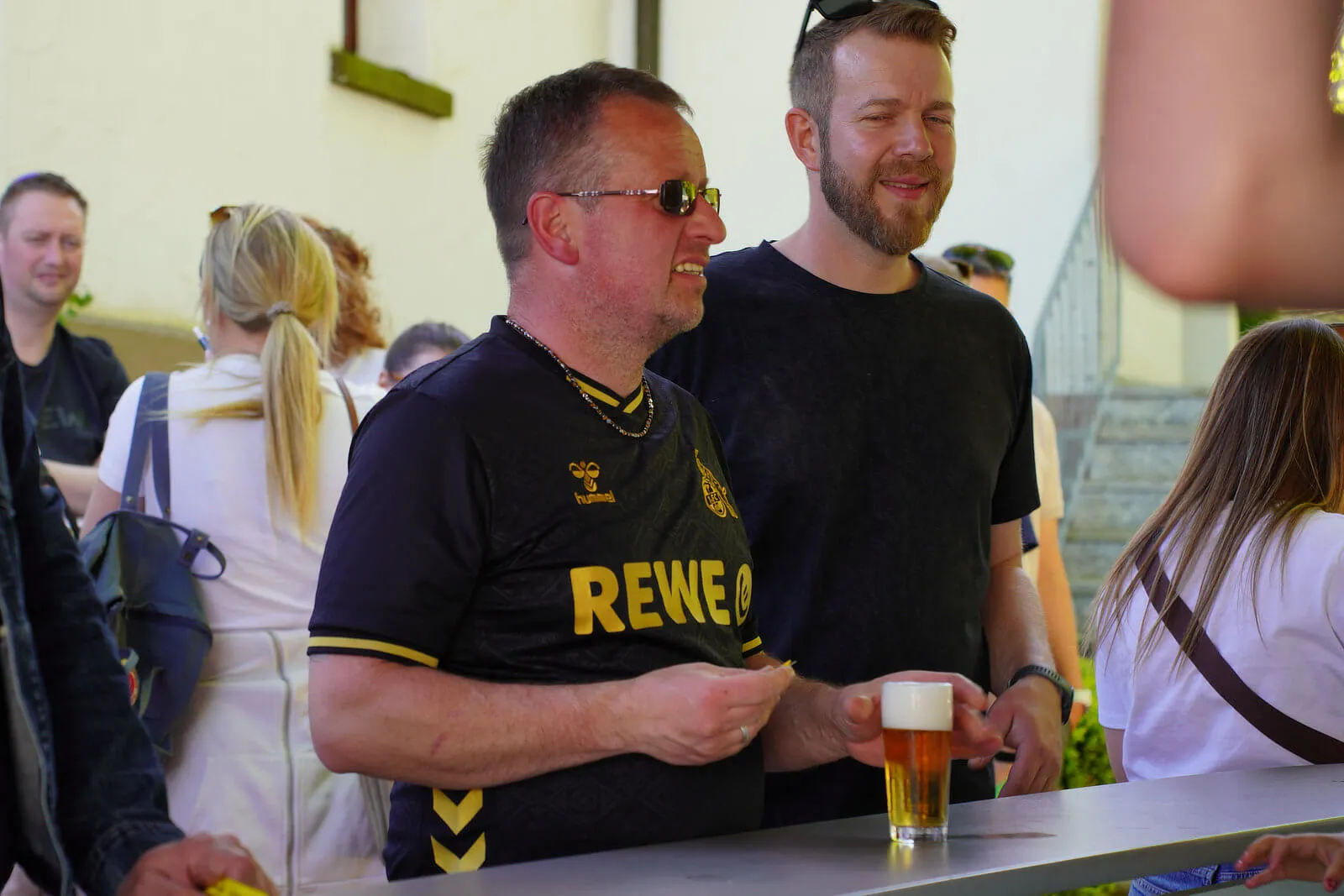Zwei lächelnde Männer stehen vor einer Theke, einer davon trägt ein Trikot mit dem Aufdruck REWE. Im Hintergrund sind weitere Menschen zu sehen.