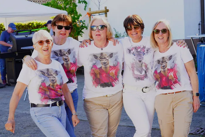 Fünf Frauen mit Sonnenbrillen posieren eng zusammen und tragen T-Shirts mit Rehen. Im Hintergrund ist ein Grill und weitere Personen zu sehen.