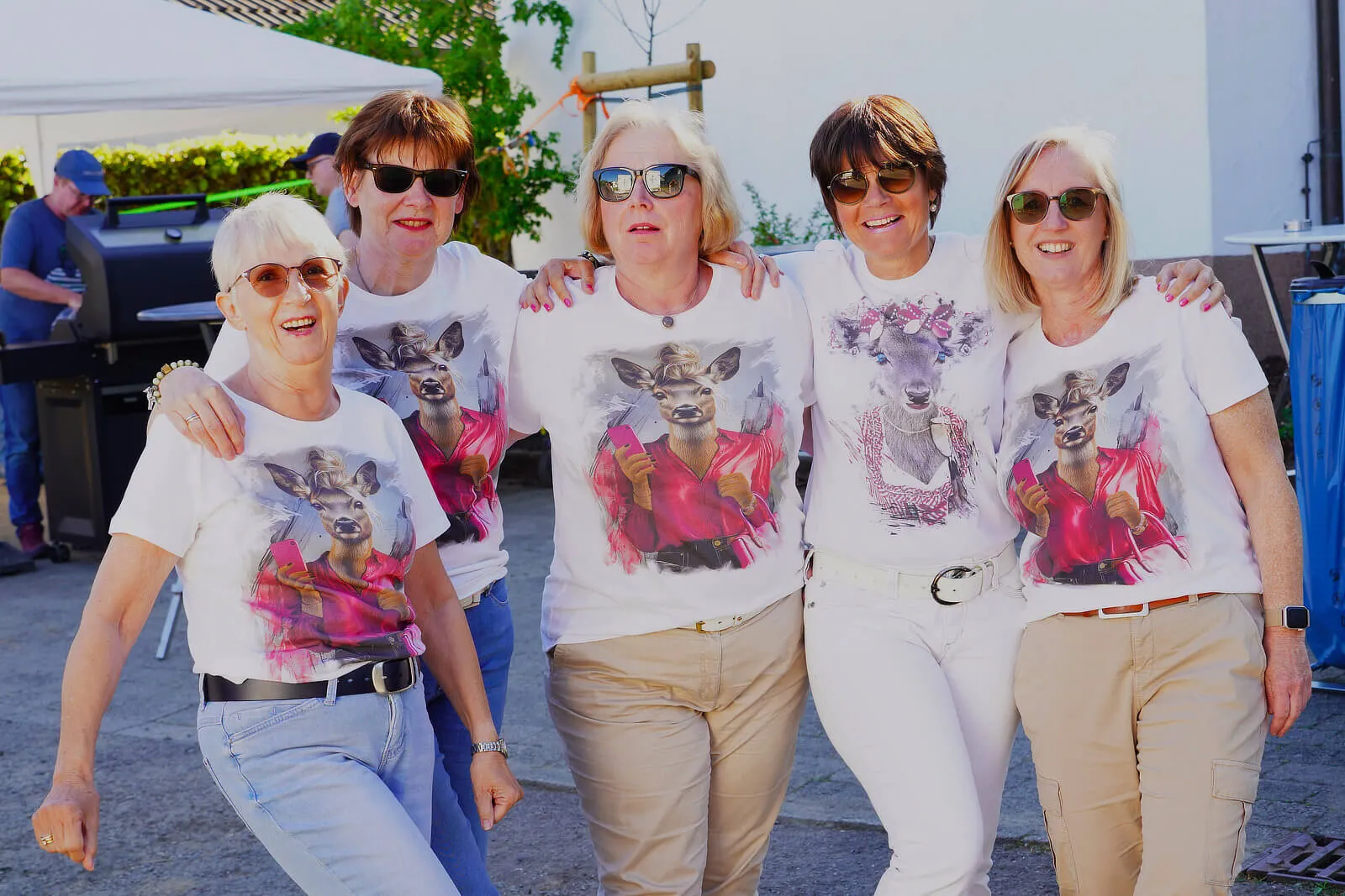 Fünf Frauen mit Sonnenbrillen posieren eng zusammen und tragen T-Shirts mit Rehen. Im Hintergrund ist ein Grill und weitere Personen zu sehen.