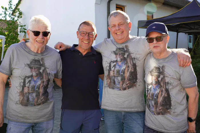 Vier lächelnde Männer stehen eng zusammen, zwei von ihnen haben Arme um die Schultern der anderen gelegt. Sie tragen T-Shirts mit einem ähnlichen Motiv und Sonnenbrillen.