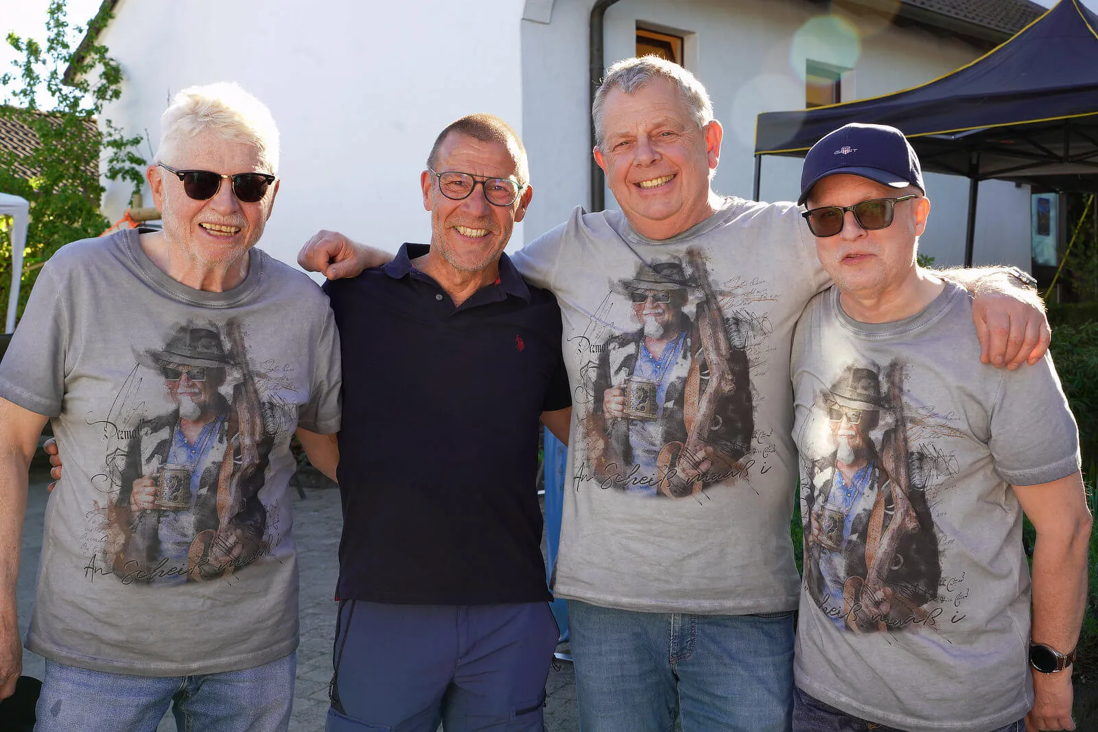 Vier lächelnde Männer stehen eng zusammen, zwei von ihnen haben Arme um die Schultern der anderen gelegt. Sie tragen T-Shirts mit einem ähnlichen Motiv und Sonnenbrillen.