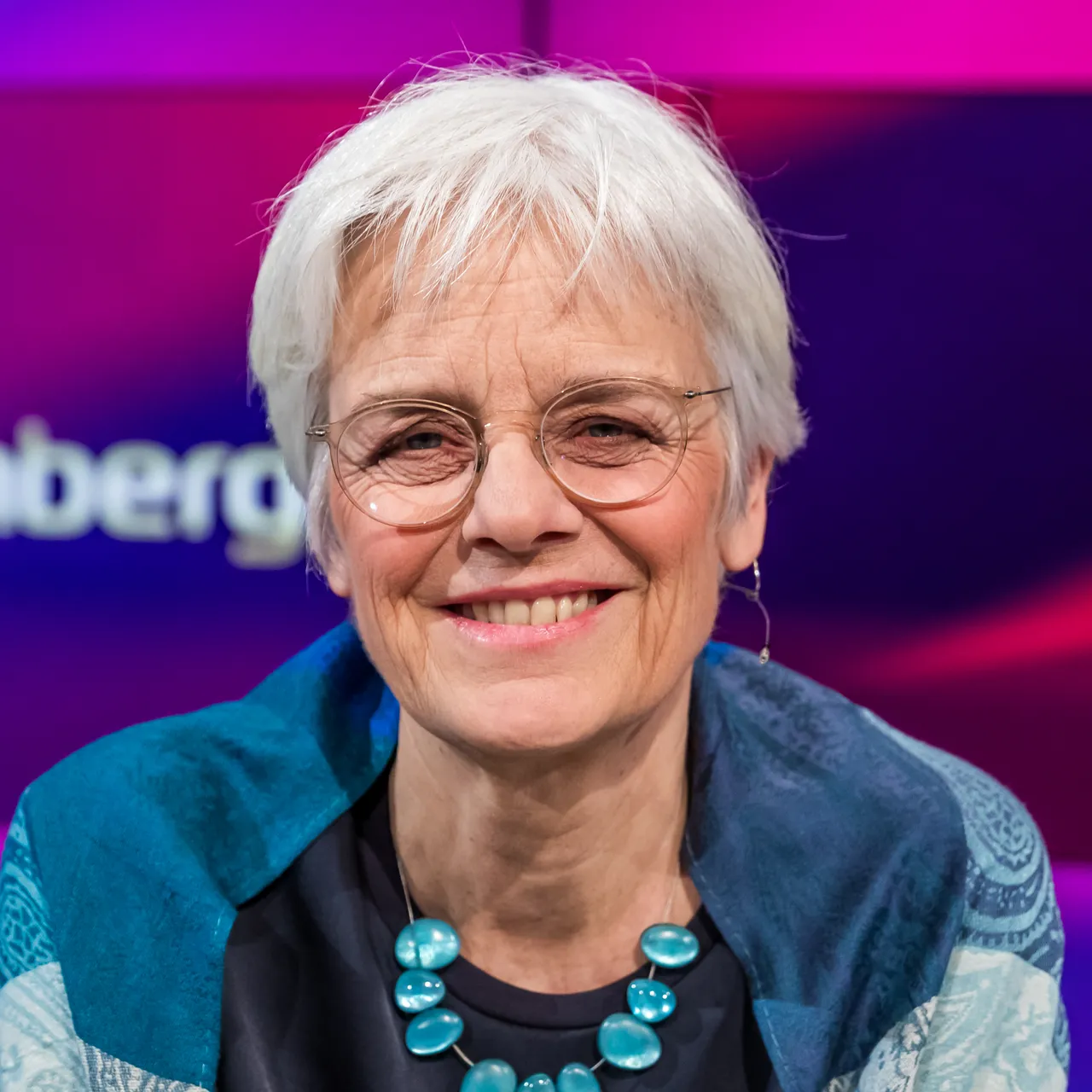 Porträt von Ulrike Herrmann mit kurzem, weißem Haar und runder Brille, lächelnd in die Kamera blickend. Sie trägt ein blaues Oberteil mit einem auffälligen türkisfarbenen Schmuckstück.