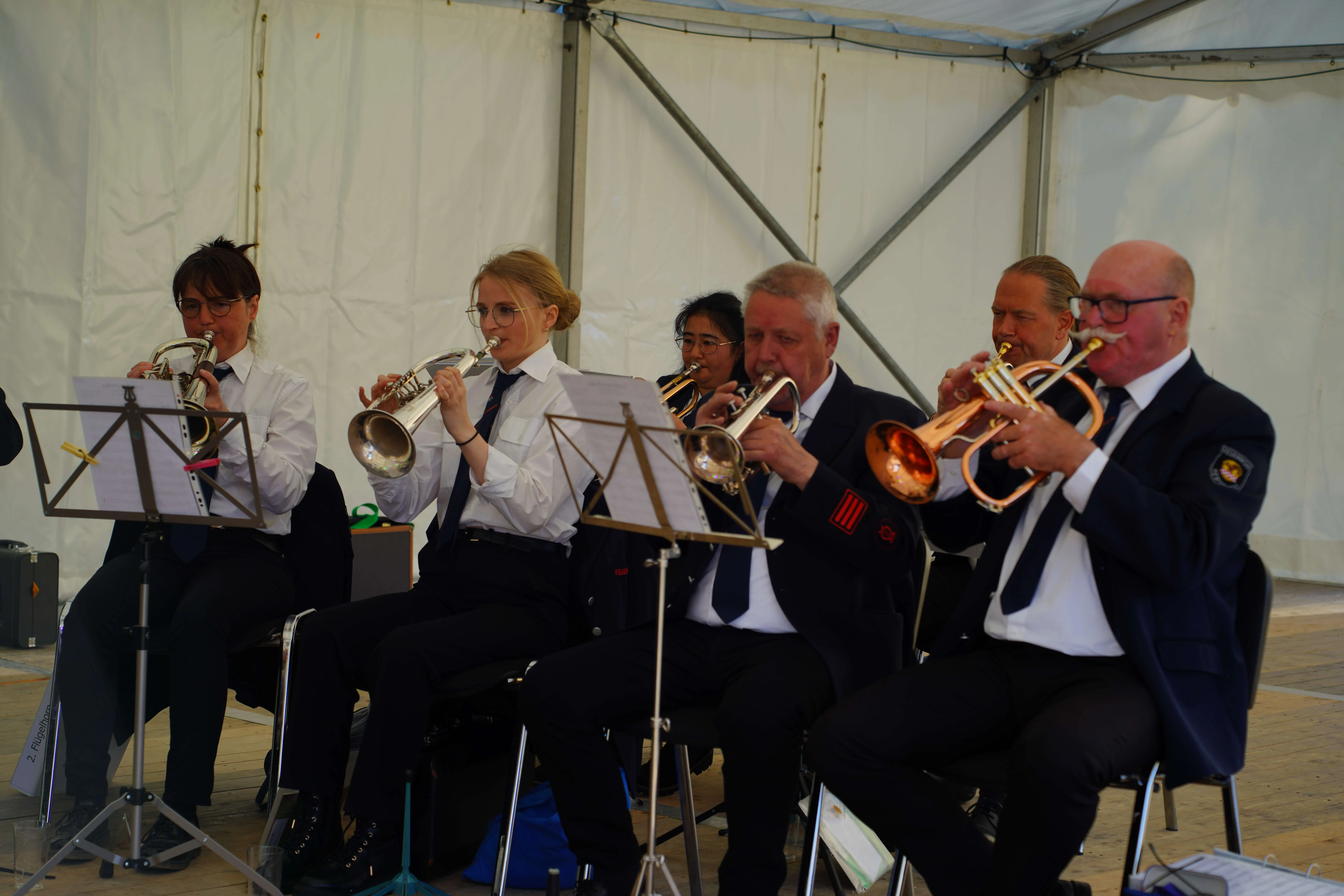 Fünf Musiker spielen Trompeten vor einem weißen Zelt, Notenständer stehen vor ihnen.