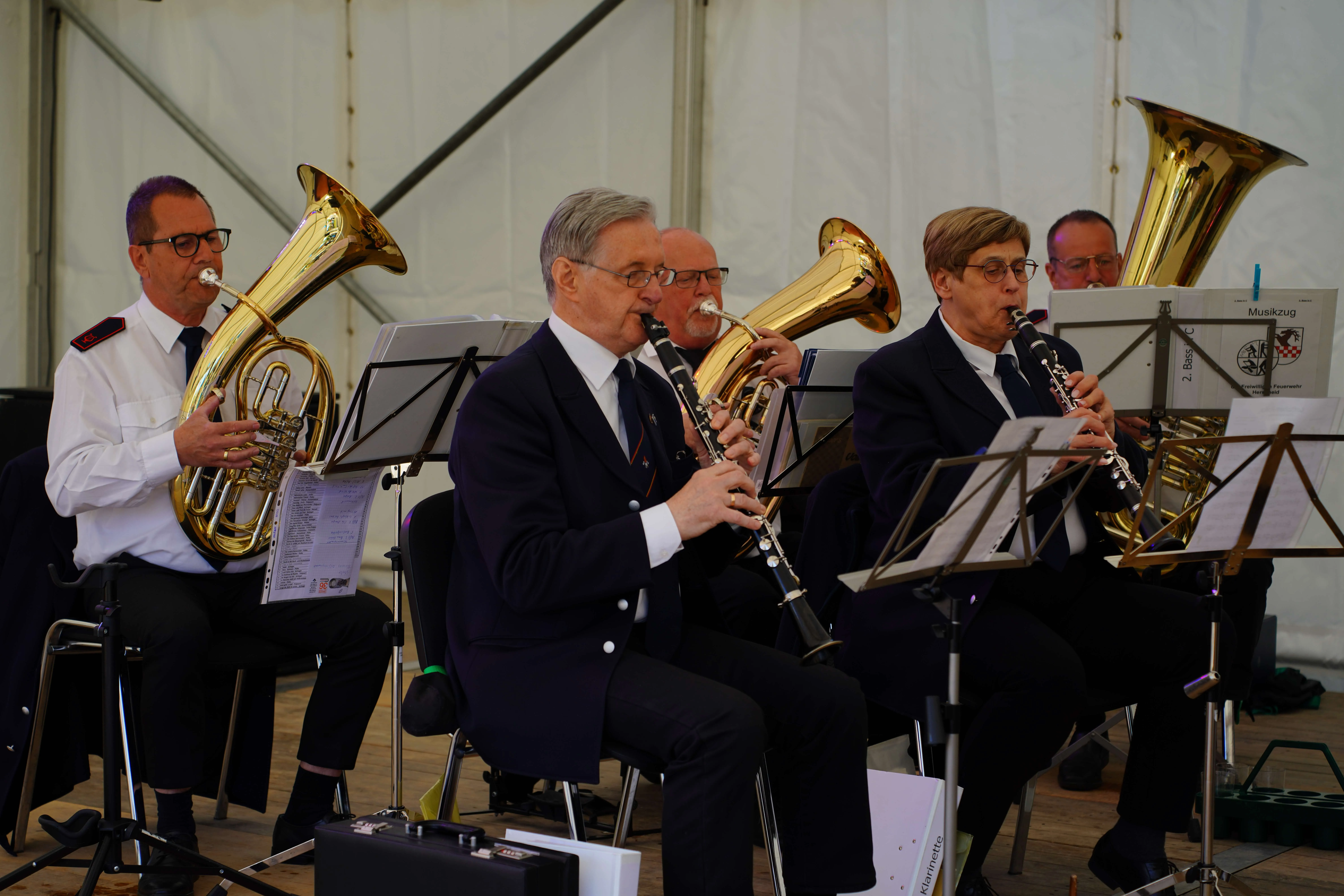 Mehrere Musiker spielen während eines Konzerts Blasinstrumente wie Posaune, Klarinette und Tuba. Notenständer stehen vor den Musikern.