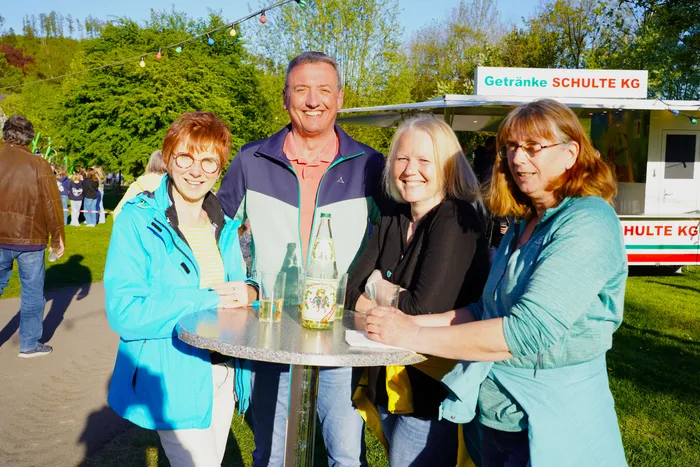Vier lächelnde Menschen stehen um einen Stehtisch mit einer Mineralwasserflasche. Im Hintergrund ein Getränkewagen und weitere Besucher des Maifests.