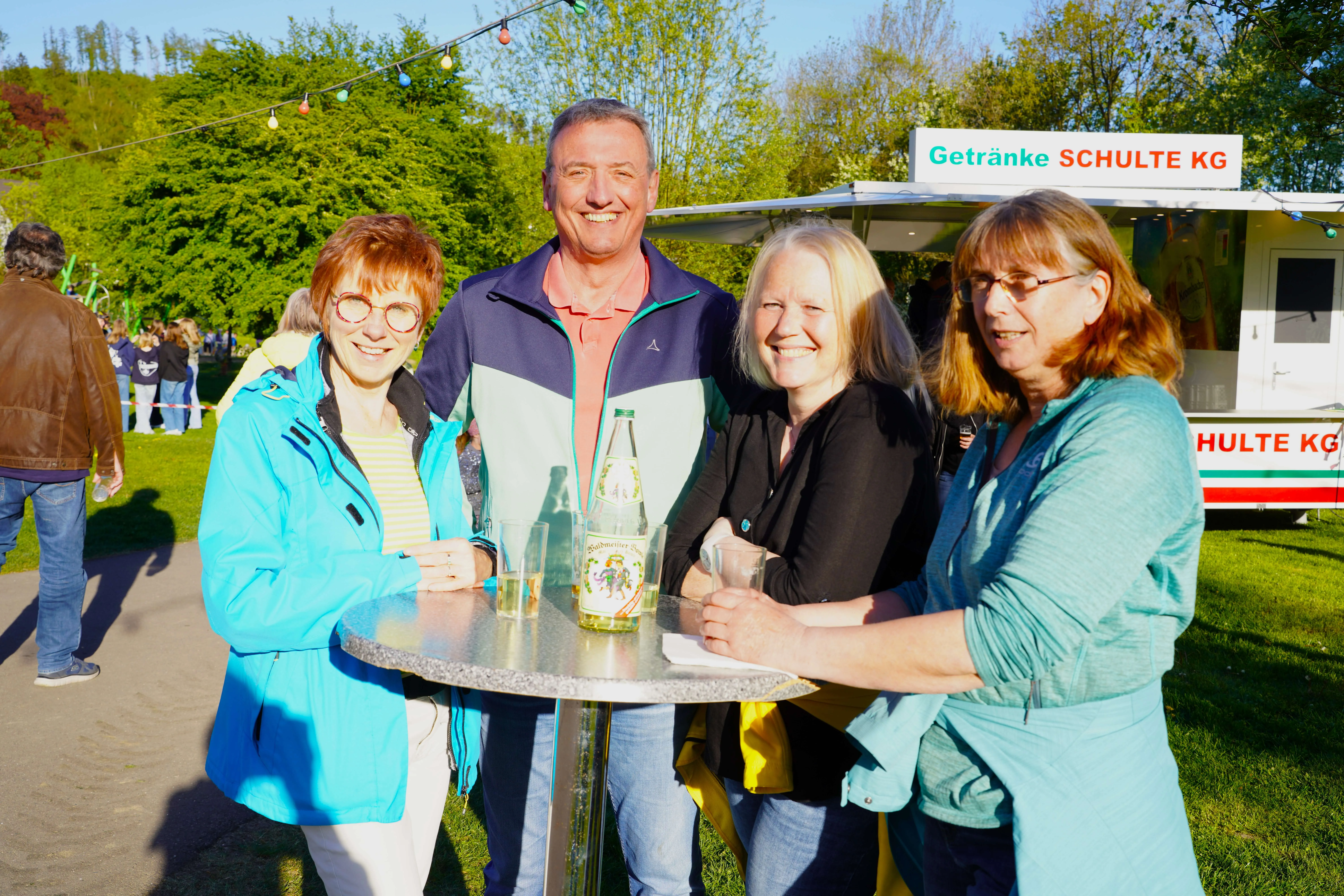 Vier lächelnde Menschen stehen um einen Stehtisch mit einer Mineralwasserflasche. Im Hintergrund ein Getränkewagen und weitere Besucher des Maifests.