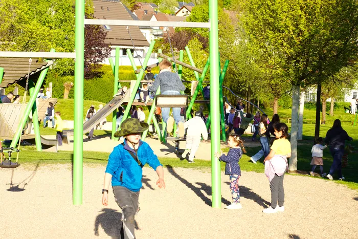 Kinder rennen und spielen auf einem Spielplatz, während im Hintergrund weitere Menschen den Tanz in den Mai feiern.