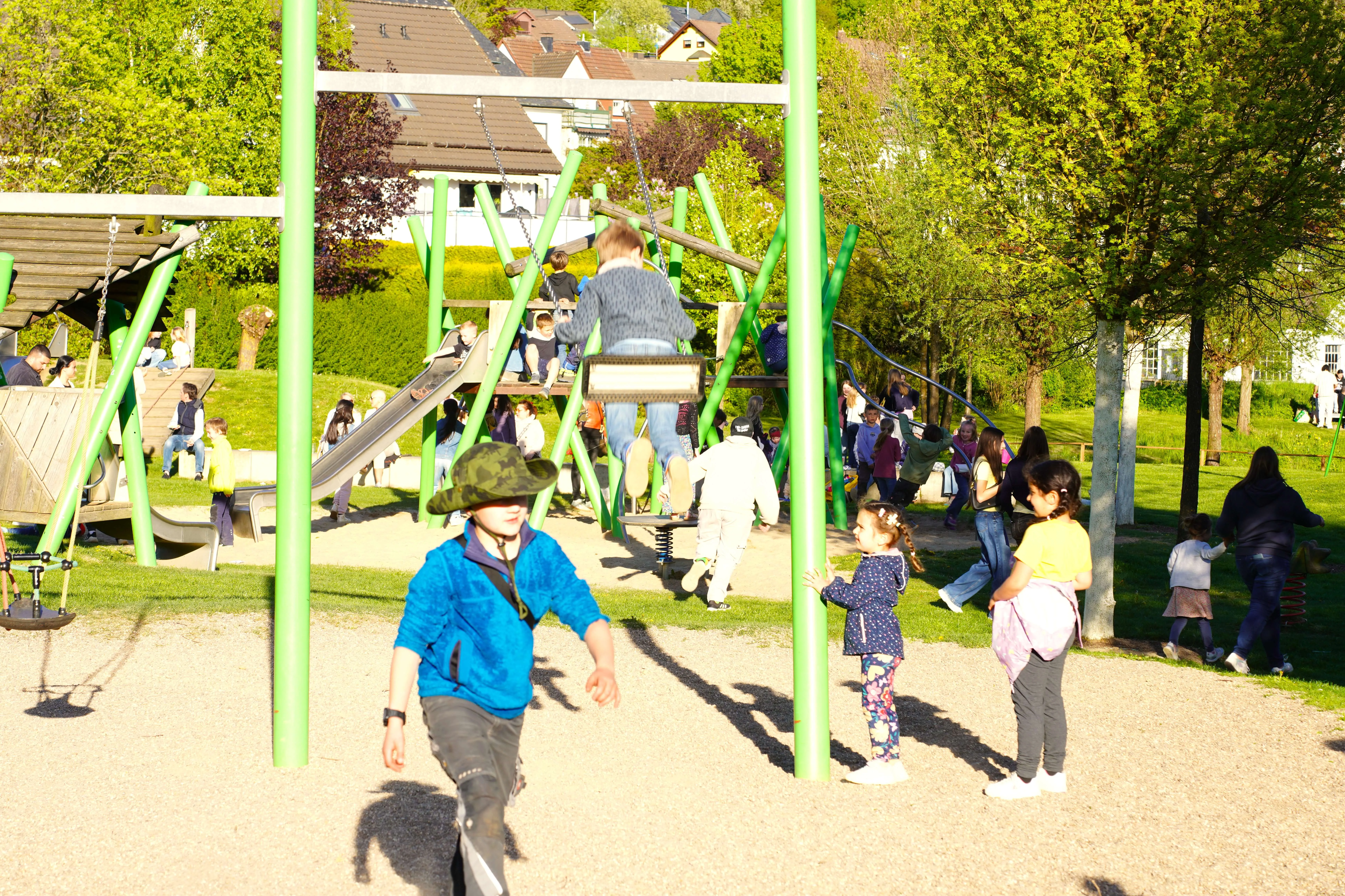 Kinder rennen und spielen auf einem Spielplatz, während im Hintergrund weitere Menschen den Tanz in den Mai feiern.