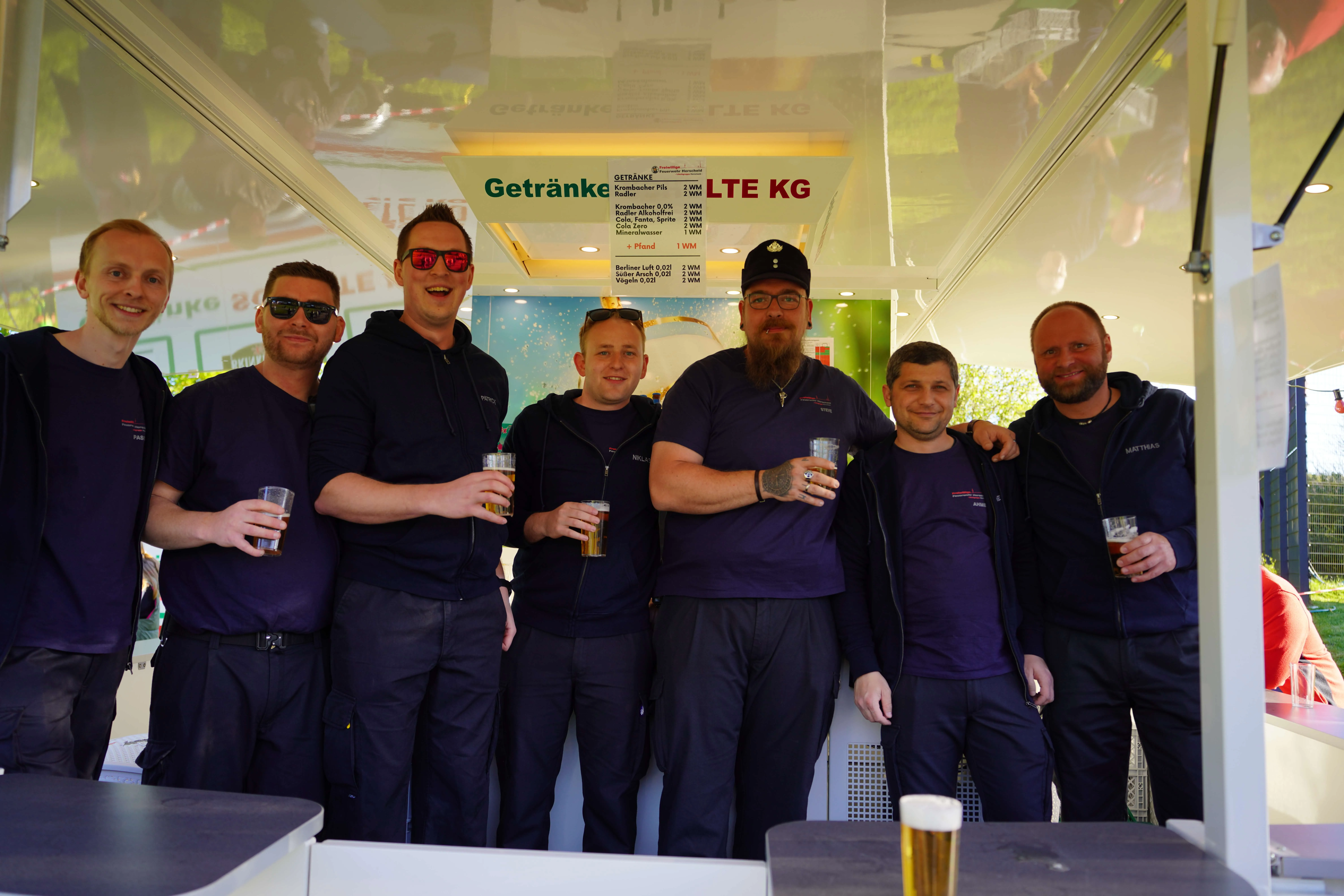 Eine Gruppe von Männern in blauen Arbeitsanzügen steht lächelnd mit Biergläsern vor einem Getränkestand mit Preisliste.