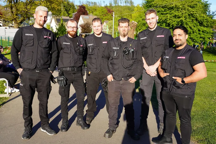 Sechs männliche Sicherheitskräfte in schwarzer Uniform und mit roten Armbinden stehen in einer Reihe vor Bäumen und Zuschauern.