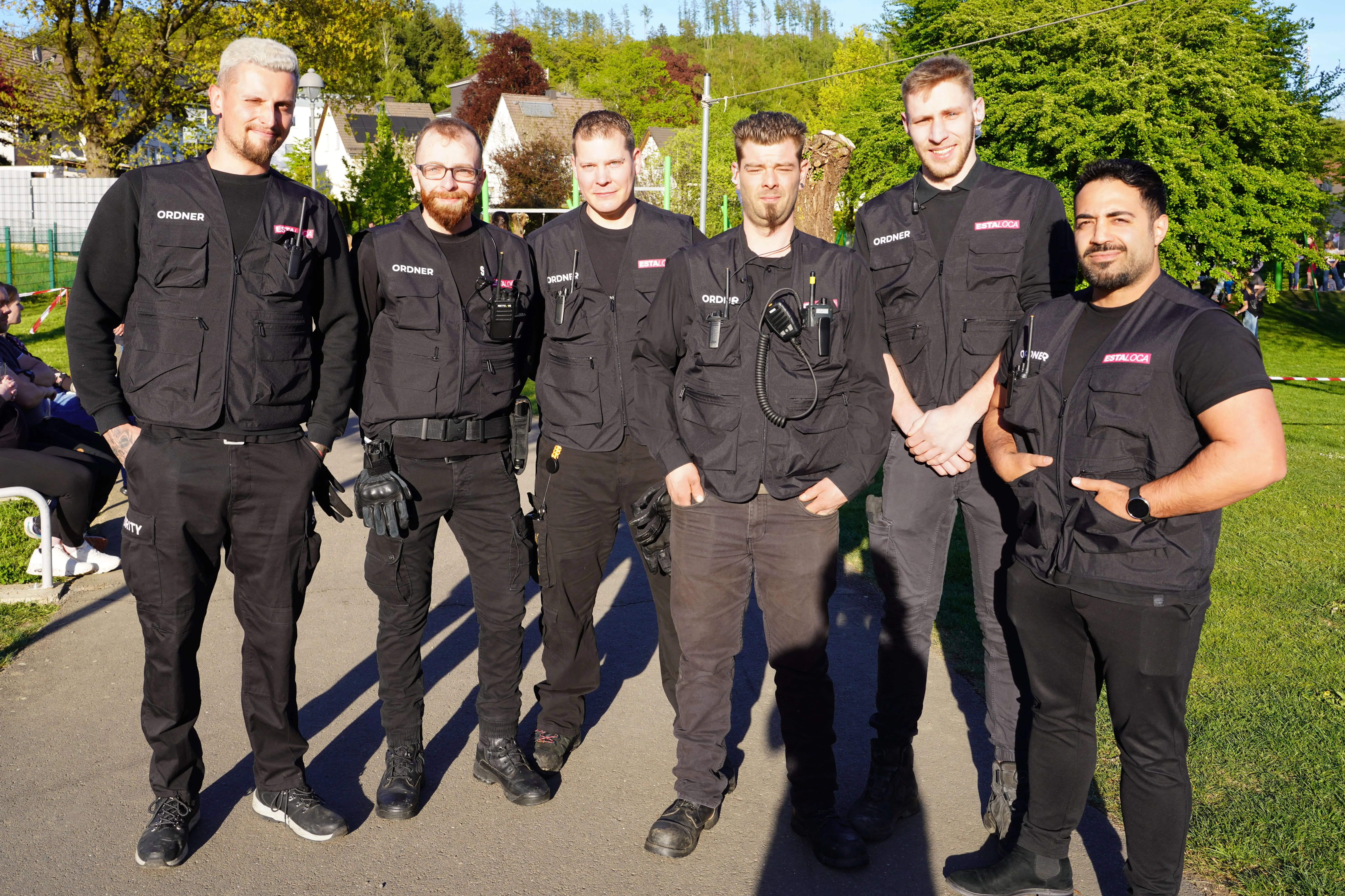 Sechs männliche Sicherheitskräfte in schwarzer Uniform und mit roten Armbinden stehen in einer Reihe vor Bäumen und Zuschauern.