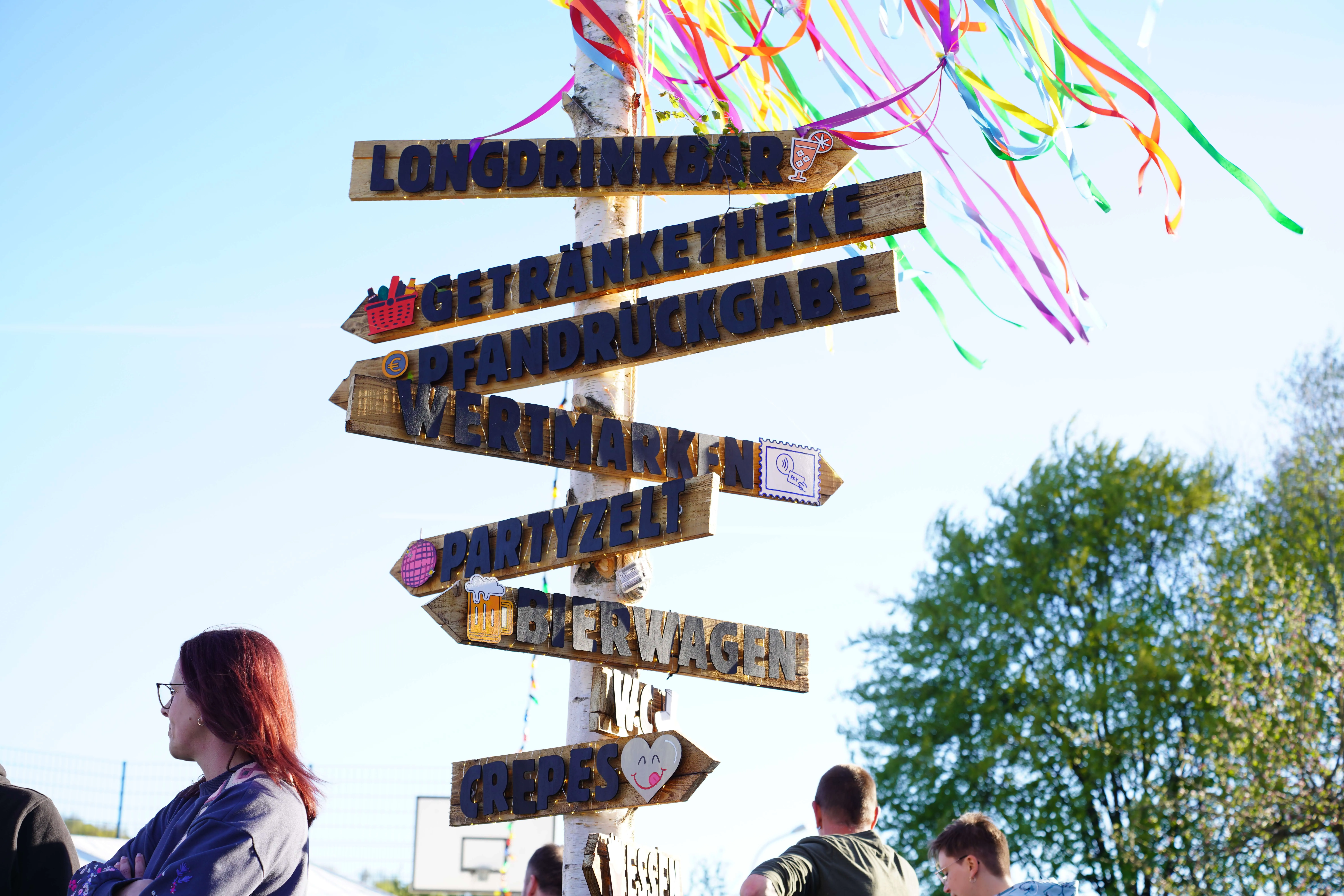 Holzschilder weisen auf verschiedene Angebote beim Maifest hin, darunter Longdrinks, Getränketheke, Pfandrückgabe, Bierwagen und Crêpes.