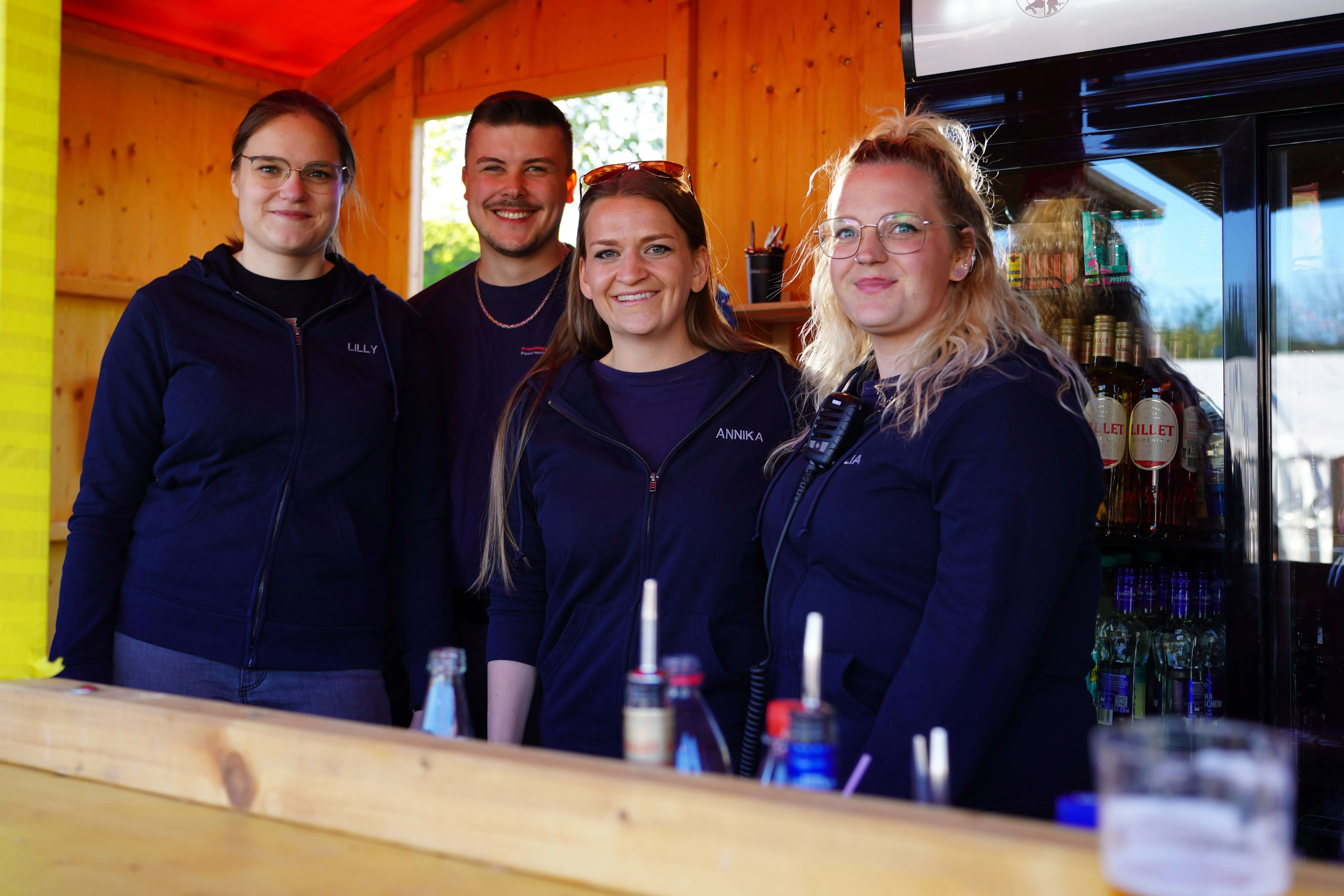 Vier lächelnde Personen stehen hinter einer Theke mit Getränken, die Namen Lilly und Annika sind auf ihren Jacken zu sehen.