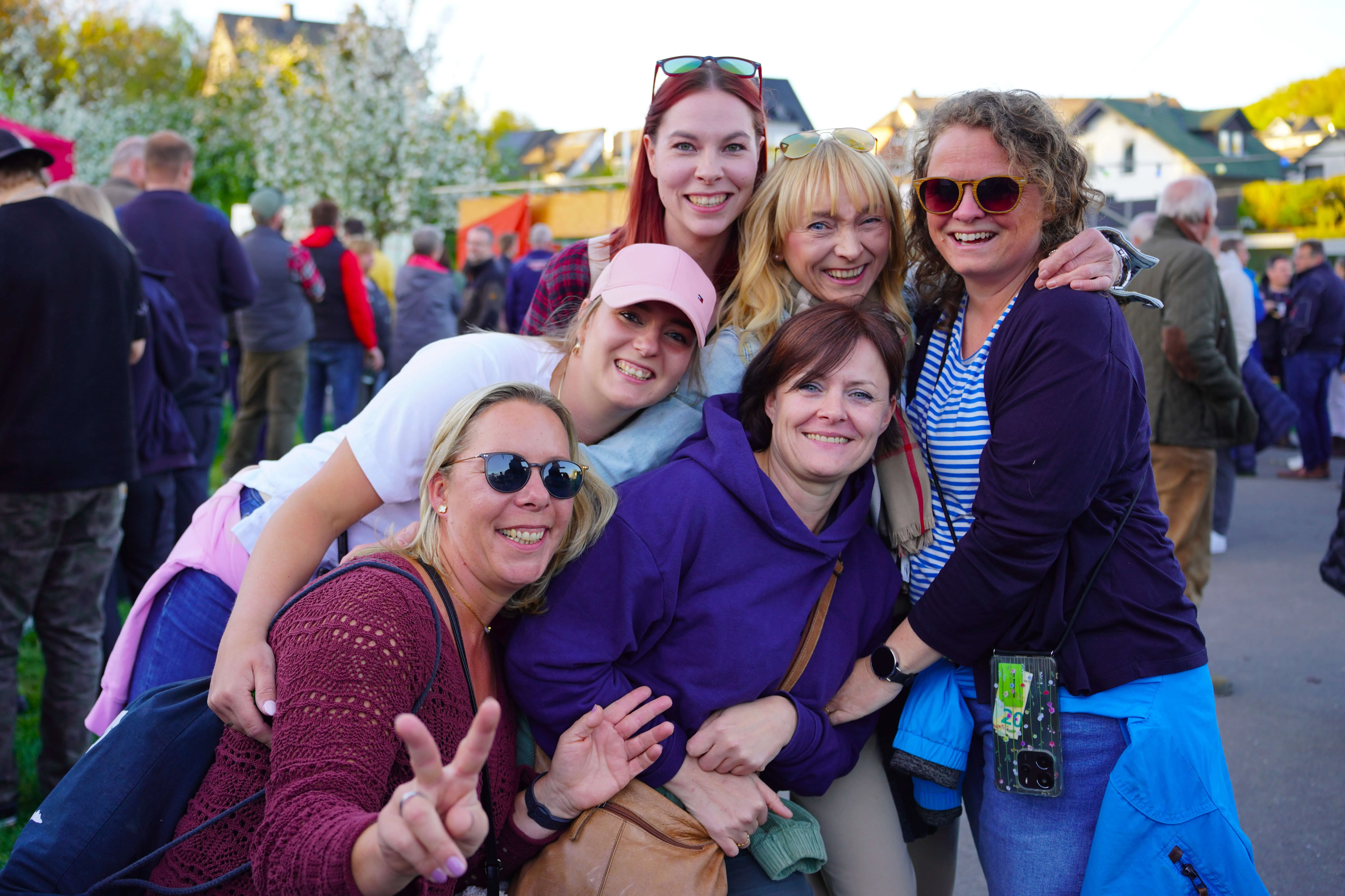 Fünf lächelnde Frauen umarmen sich eng vor einer Menschenmenge bei einer Frühlingsveranstaltung im Freien.