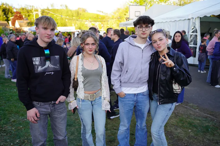 Vier junge Leute posieren im Freien vor einer Menschenmenge, die sich auf einem Festplatz versammelt hat. Im Hintergrund sind Zelte und Lichterketten zu sehen.