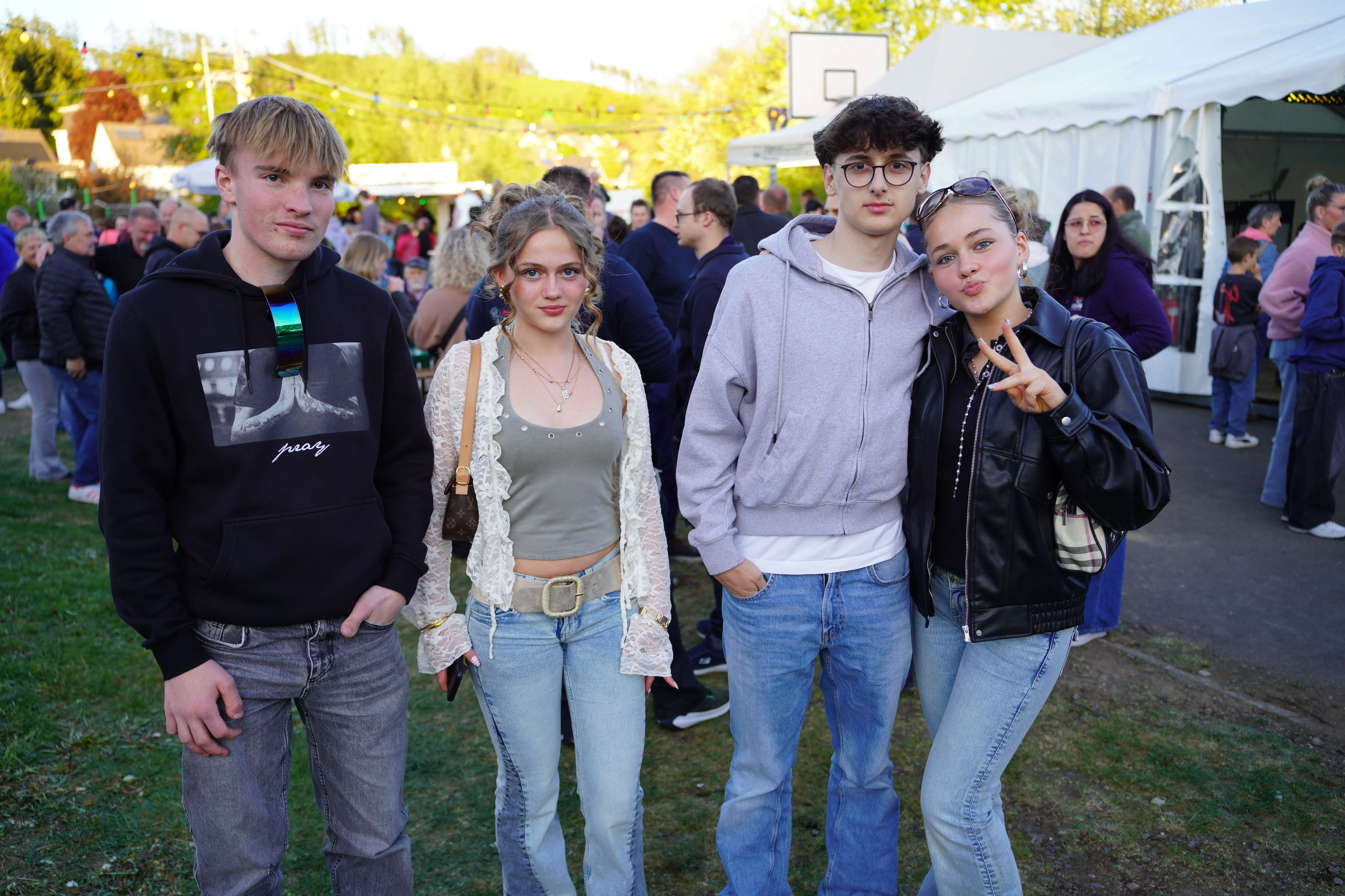 Vier junge Leute posieren im Freien vor einer Menschenmenge, die sich auf einem Festplatz versammelt hat. Im Hintergrund sind Zelte und Lichterketten zu sehen.