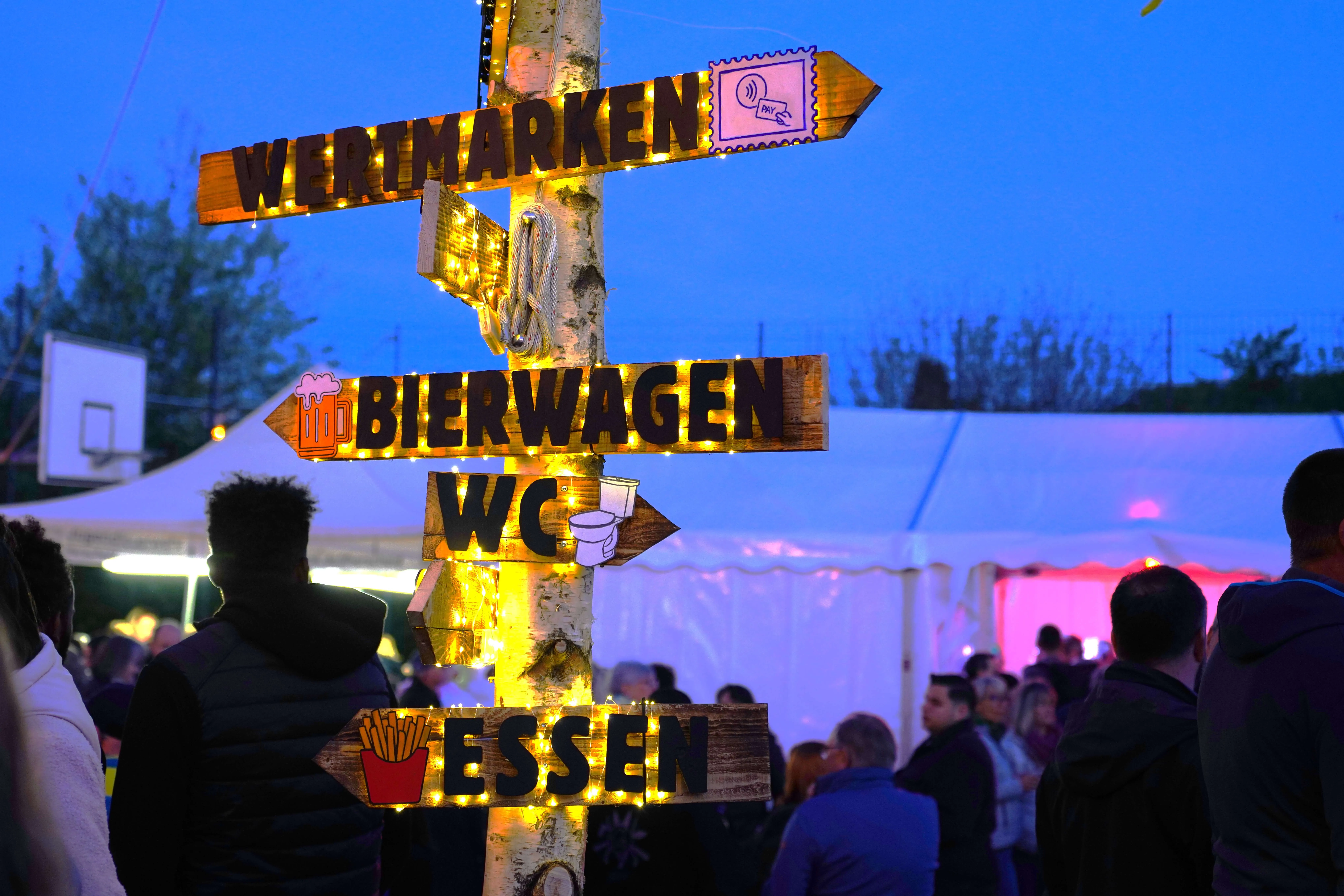 Holzschilder weisen auf ‚Wertmarken‘, ‚Bierwagen‘, ‚WC‘ und ‚Essen‘. Im Hintergrund sind Menschen und Zelte zu sehen.