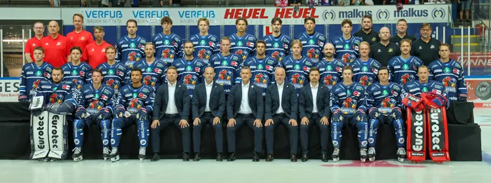 Mannschaftsfoto der Iserlohn Roosters mit Spielern in blauen Trikots und Trainern in Anzügen auf dem Eis.