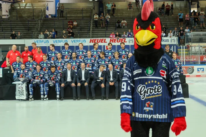 Das Maskottchen Icey steht im Vordergrund vor der Mannschaft der Iserlohn Roosters, die in blauen Trikots auf dem Eis posiert.