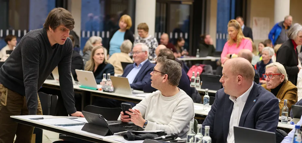 Manuel Huff steht vor Karsten Meininghaus und Fabian Tigges an einem Konferenztisch, während andere Teilnehmer im Hintergrund sitzen und zuhören.