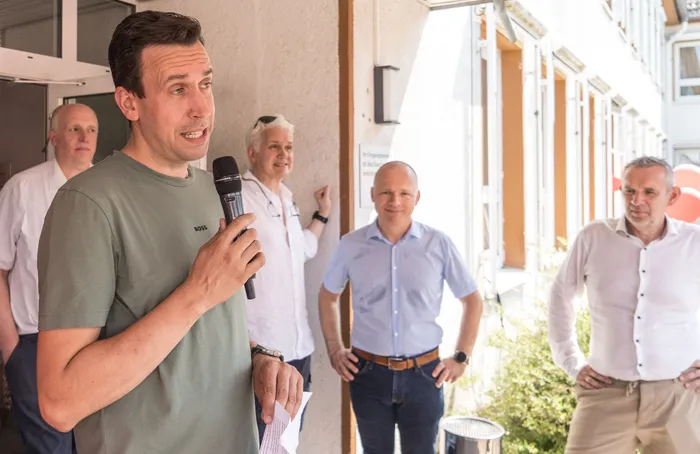 Ein Mann spricht in ein Mikrofon, umgeben von weiteren Personen vor einer hellen Fassade. Im Hintergrund sind weitere Menschen und ein Gebäude mit Säulen zu sehen.