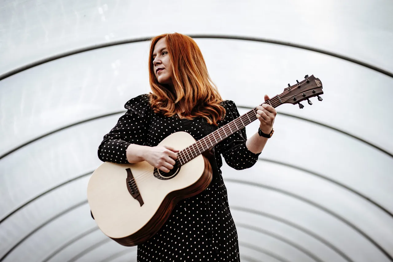 Marie-Luise Gunst spielt Gitarre vor einem Hintergrund aus weißen, geschwungenen Linien.